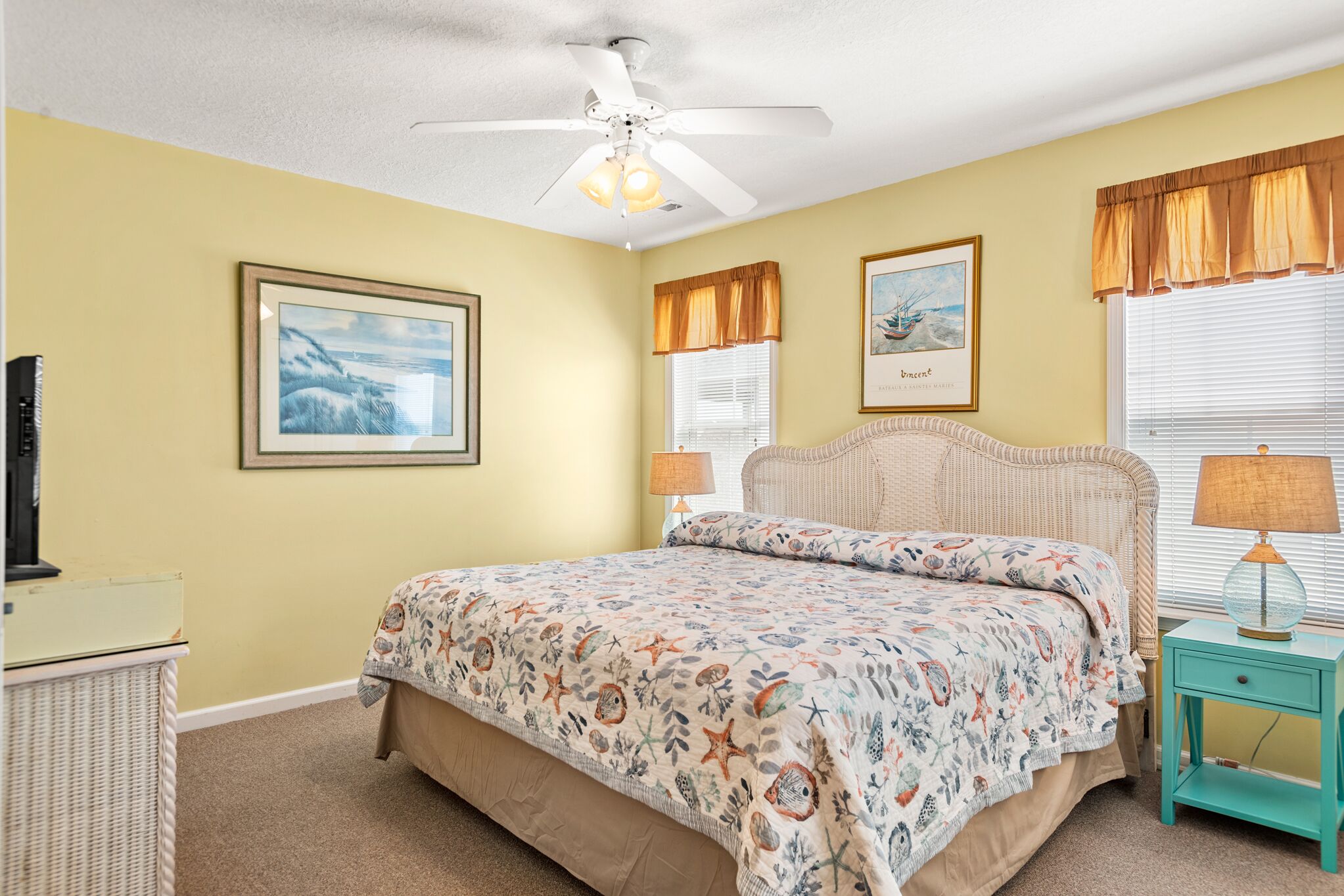 A bedroom with a bed, a ceiling fan, a nightstand, a lamp, and framed pictures on the wall.