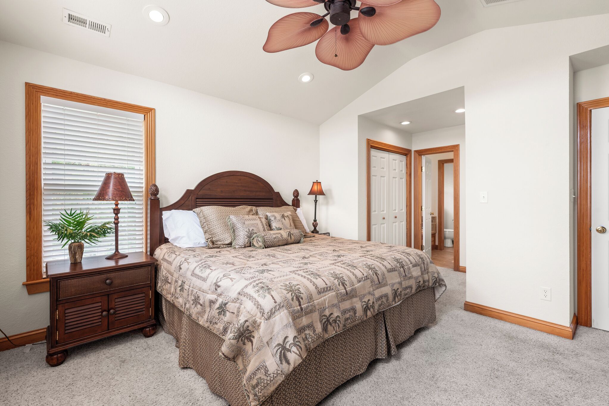 A bedroom with a large bed, a ceiling fan, a nightstand with a lamp, and a window with blinds.