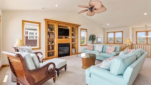 A spacious living room with a fireplace, wooden entertainment center, and a large window. - Jockeys Ridge 385: Oceanside, Private Pool, Spa (Nags Head)