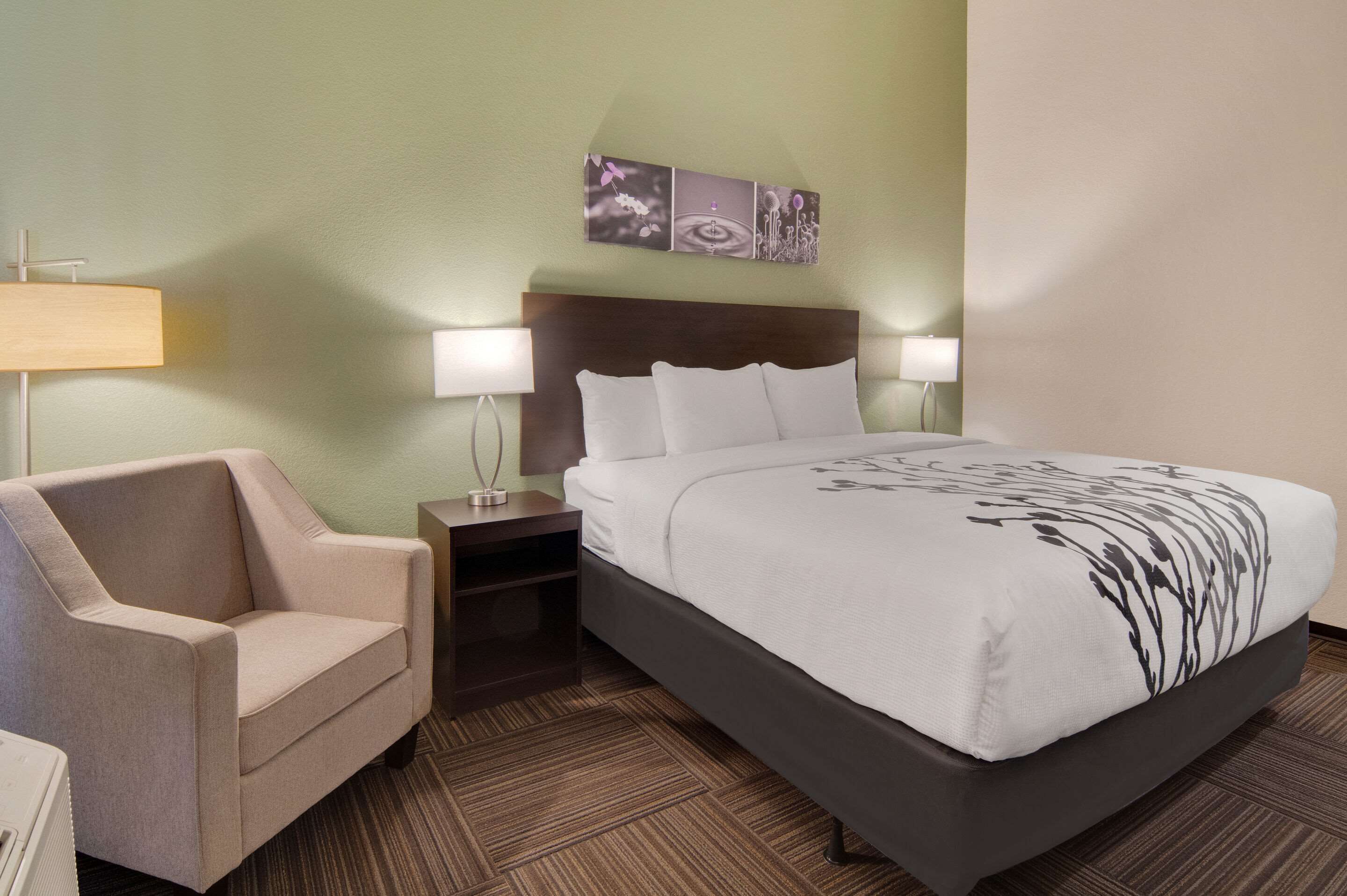 Habitación de hotel con cama, mesita de noche, lámpara, silla y una obra de arte en la pared.