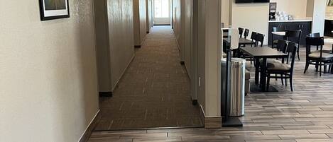 A hotel corridor with a wooden floor, a framed picture on the wall, and a seating area with tables and chairs.