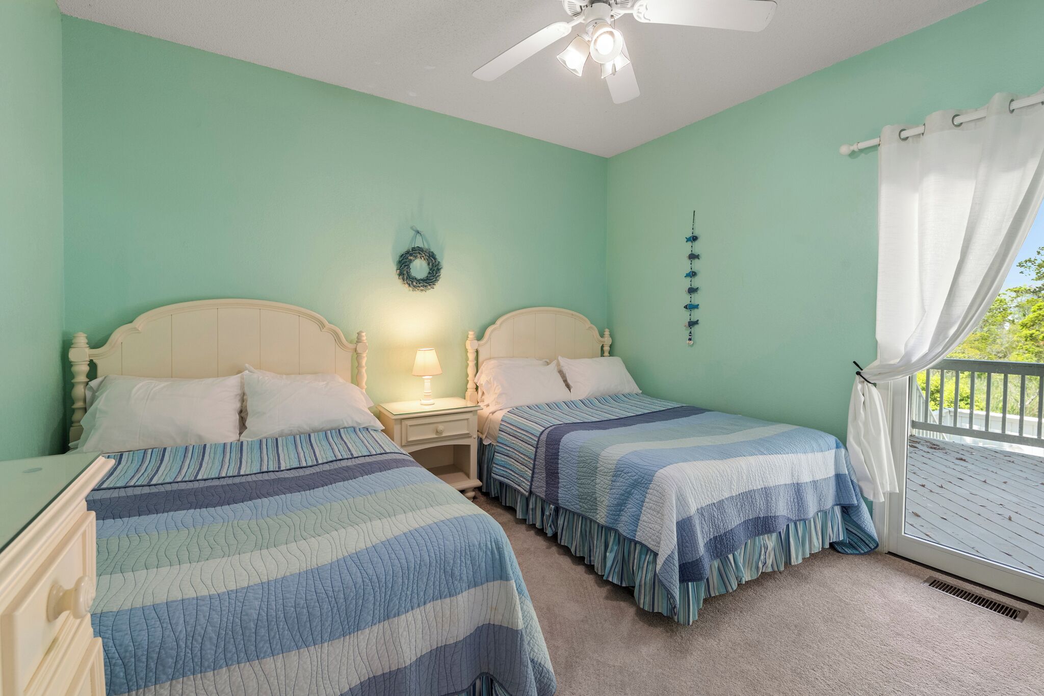 A bedroom with two beds, a ceiling fan, a nightstand with a lamp, a window with curtains, and a wall decoration.