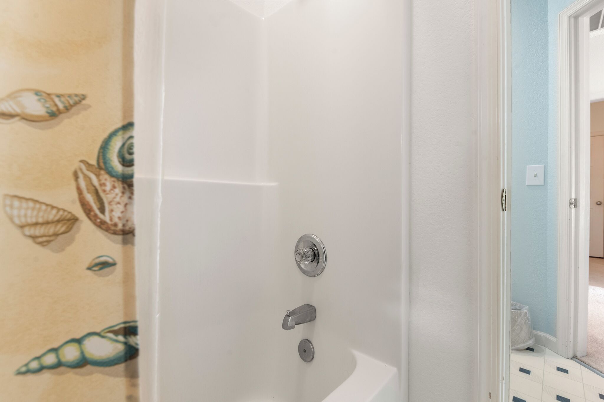 Une salle de bain avec une baignoire blanche, un rideau de douche orné d’un motif de coquillages et une porte donnant sur une autre pièce.