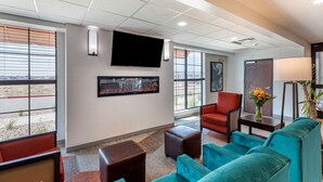 A hotel lobby with a flat-screen TV mounted on the wall, a red armchair, teal sofas, and a small table with a vase of flowers.