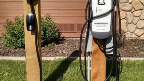Two electric vehicle charging stations mounted on wooden posts in front of a building with a wooden facade and a stone wall.