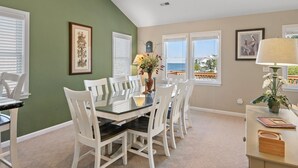 A dining area with a glass table surrounded by white chairs, framed artwork on the walls, and a view of the ocean through the windows. - Birdie: Private pool, Spa, Golf discounts (Corolla)