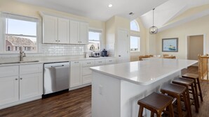 A modern kitchen with white cabinetry, a large island with bar stools, and a view of the neighborhood. - Soundset : Soundview Private Spa, Golf, Share Pool (Nags Head)