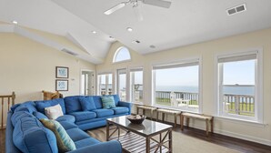 A spacious living room with a blue sectional sofa, a wooden coffee table, and a view of the ocean through large windows. - Soundset : Soundview Private Spa, Golf, Share Pool (Nags Head)