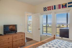 Une chambre dotée d’une télévision, d’une commode en bois et d’un lit recouvert d’une couvre-lit à motifs. Deux fenêtres offrent une vue sur une terrasse et l’océan.