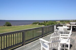 Une terrasse dotée de chaises Adirondack blanches et d’un barbecue, offrant une vue sur une pelouse et la mer.