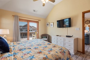 A bedroom with a large bed, a television, a dresser, and a view of a house through a sliding door. - Seaduction: Oceanside, 6-bedrooms, Private Pool & Hot Tub, 3 lots from the ocean (Corolla)