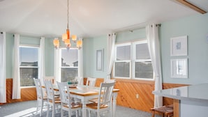 Een eetruimte met een houten tafel en witte stoelen, een hanglamp en grote ramen met witte gordijnen.