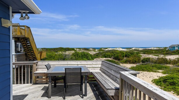 Een houten terras met een tafel en stoelen, met uitzicht op een strand met duinen en een huis in de verte.