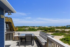 A wooden deck with a table and chairs, overlooking a beach with dunes and a distant house. - Lion on the Beach: Ocean Views, 4-bedroom home, short walk to the beach (Corolla)