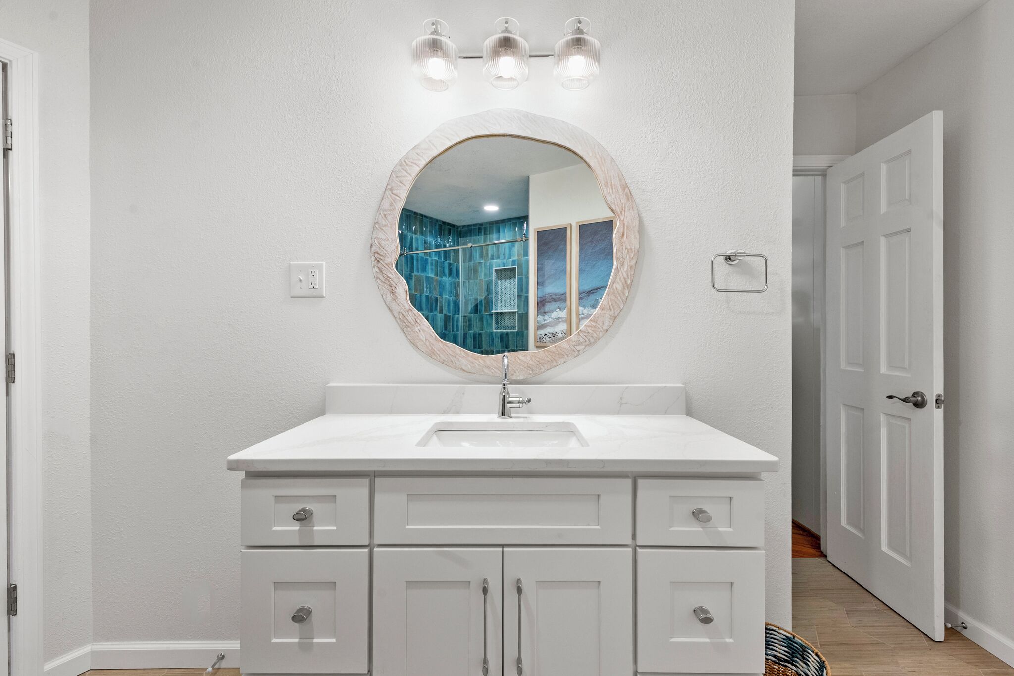 A bathroom with a white vanity, a round mirror with a wooden frame, and a blue-tiled shower area visible in the reflection.