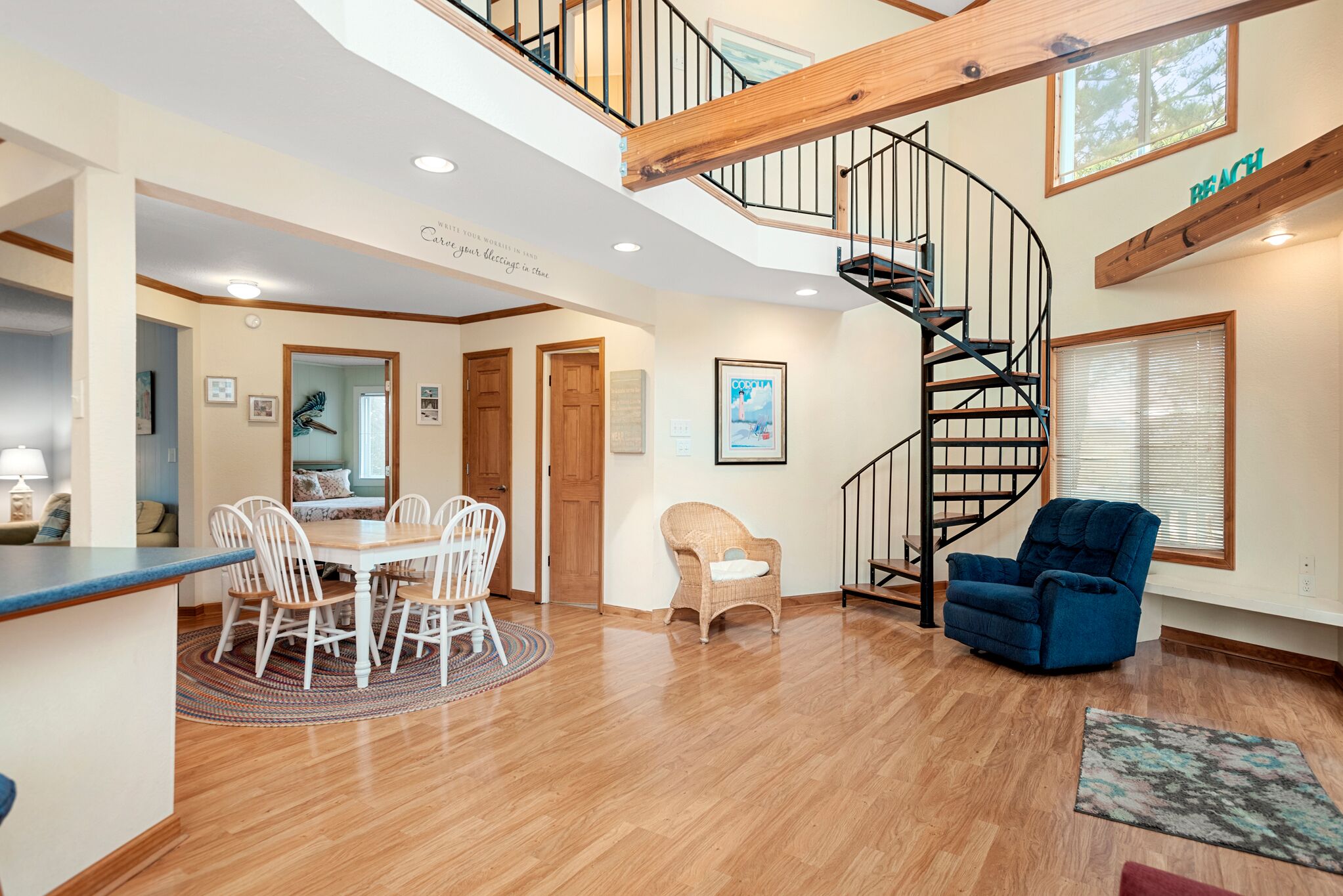 A spacious living area with a wooden floor, a dining table with chairs, a blue armchair, a wicker chair, and a spiral staircase.