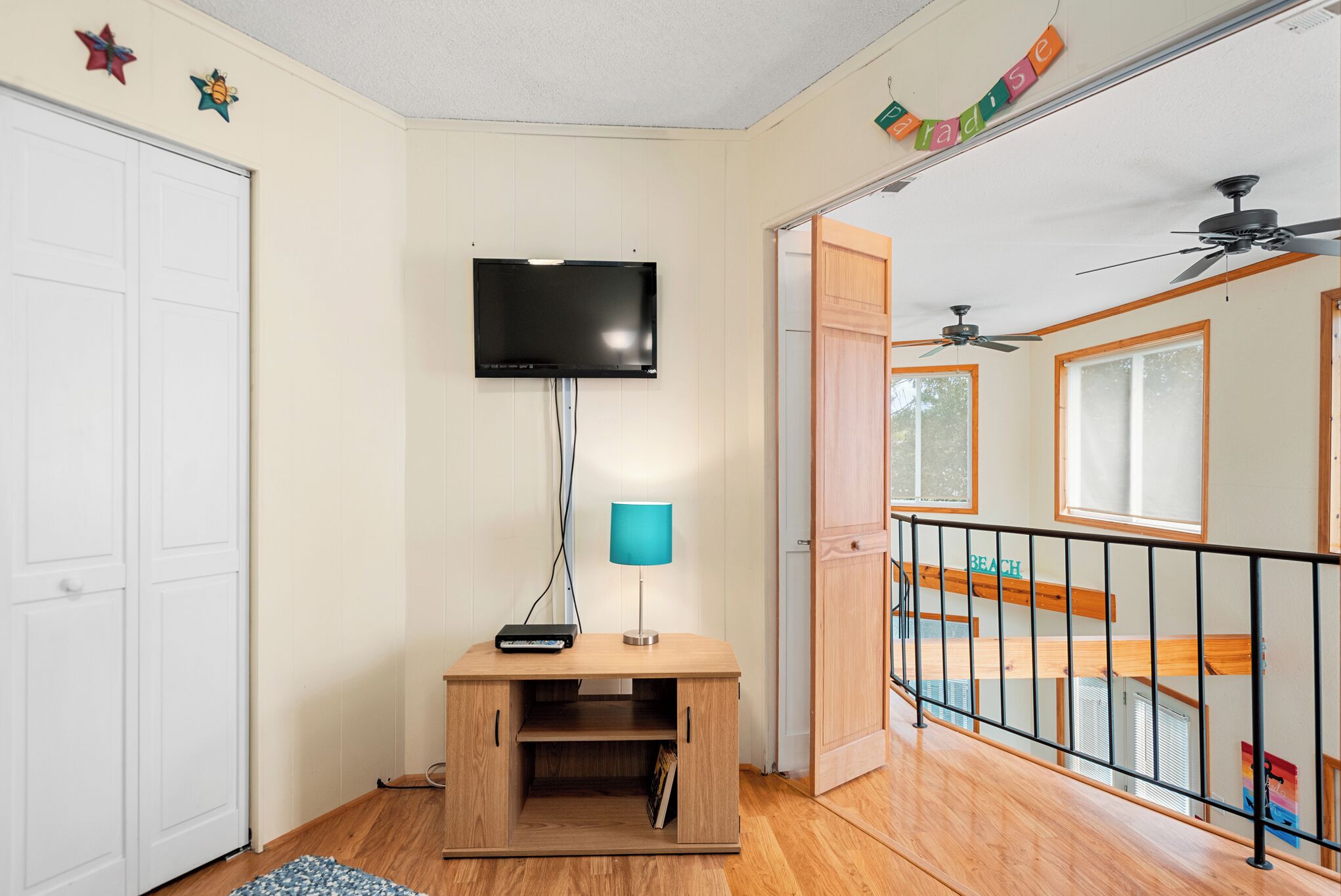 A room with a TV mounted on the wall, a wooden cabinet, a blue lamp, and a balcony with a railing.
