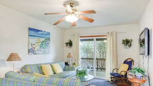 Ein Wohnzimmer mit einem gestreiften Sofa, einem Deckenventilator, einem Gemälde von Strandstühlen und Blick durch eine Schiebetür.