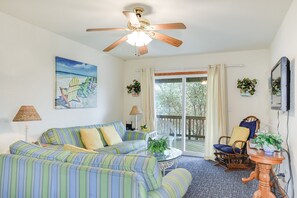 Ein Wohnzimmer mit einem gestreiften Sofa, einem Deckenventilator, einem Gemälde von Strandstühlen und Blick durch eine Schiebetür.