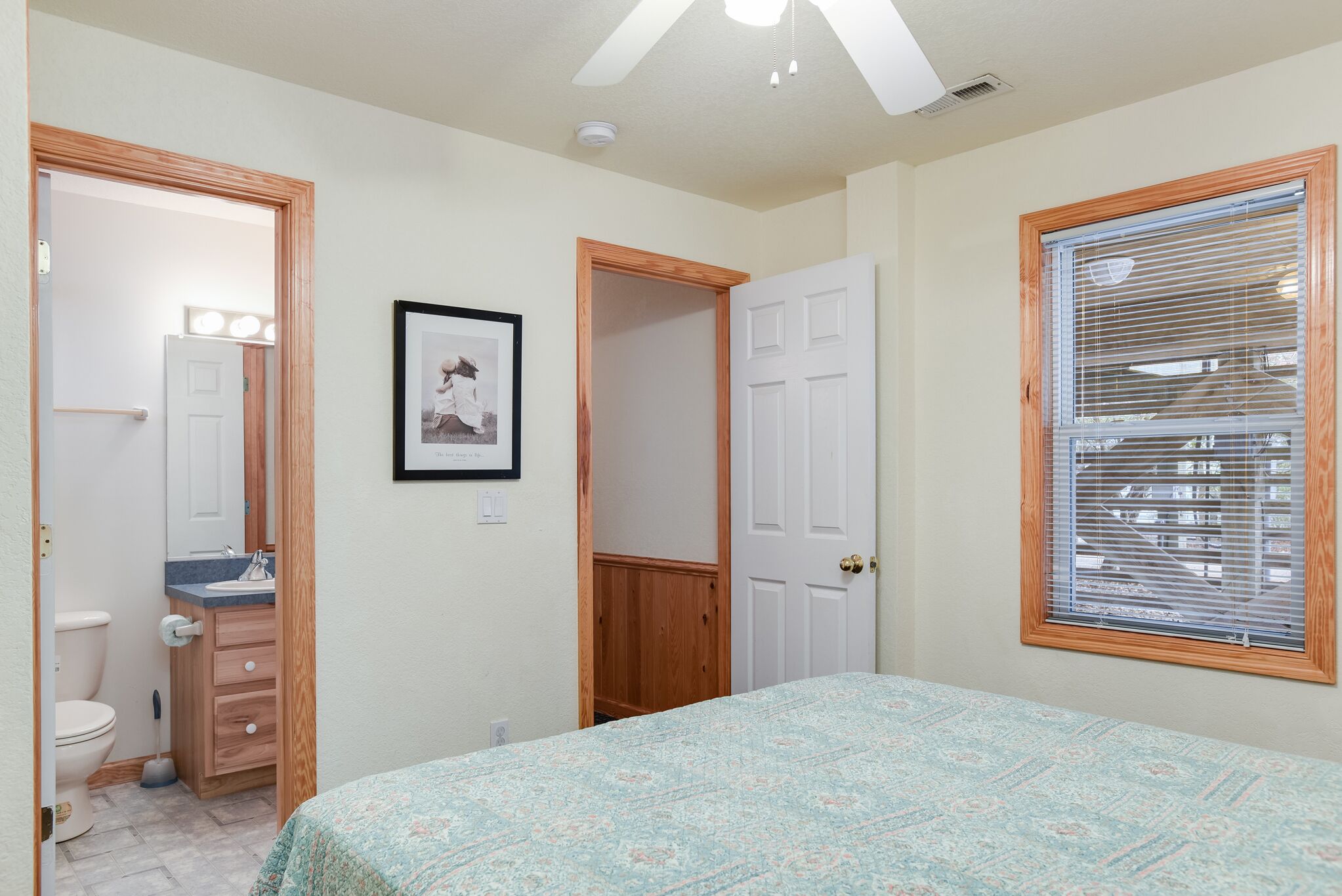 Une chambre à coucher avec un lit, une salle de bain avec des toilettes, un lavabo et un miroir. Il y a une porte qui donne sur une autre pièce et une fenêtre avec des stores.