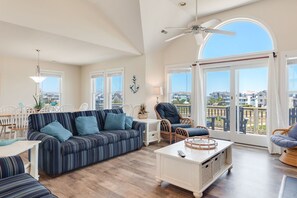 A spacious living room with a large sofa, a coffee table, and wicker chairs. There is a dining area with a table and chairs, and large windows offering a view of the city.