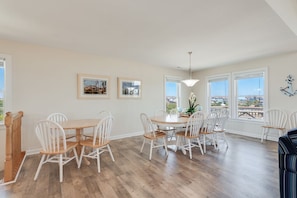Uma sala de jantar com mesa de madeira e cadeiras brancas, quadros nas paredes e janelas amplas com vista.