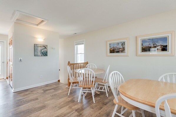 A dining area with a wooden table and white chairs, a white wall with framed pictures, and a wooden floor.