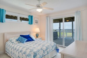 A bedroom with a bed, a nightstand, a lamp, and a view of a deck and ocean.