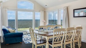 A dining area with a table and chairs, large windows with a view of the water, a blue sofa, and a chandelier. - Sunsets Forever: Soundfront, Private Pool, Hot Tub (Nags Head)