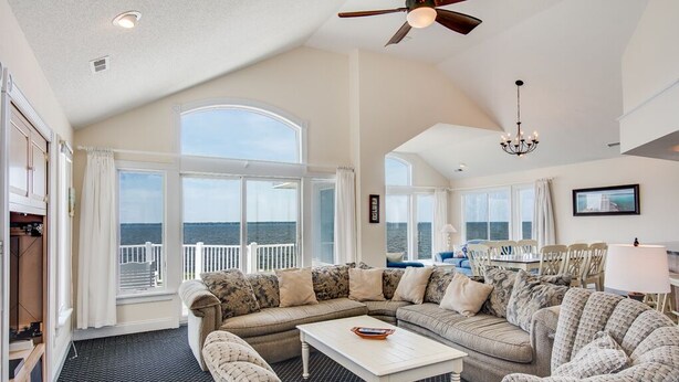 A spacious living room with a large window overlooking the sea, a ceiling fan, and a chandelier. - Sunsets Forever: Soundfront, Private Pool, Hot Tub (Nags Head)