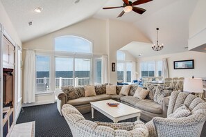 A spacious living room with a large window overlooking the sea, a ceiling fan, and a chandelier. - Sunsets Forever: Soundfront, Private Pool, Hot Tub (Nags Head)