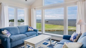 A living room with a blue sofa, white coffee table, and large windows overlooking a body of water. - Sunsets Forever: Soundfront, Private Pool, Hot Tub (Nags Head)