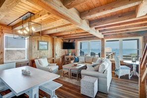 Un intérieur de chalet chaleureux avec des poutres en bois, un coin repas, des sièges et de grandes fenêtres donnant sur l’océan.