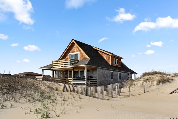 Ein Strandhaus mit Holzfassade, einem Balkon und einem Sandstrand im Hintergrund.