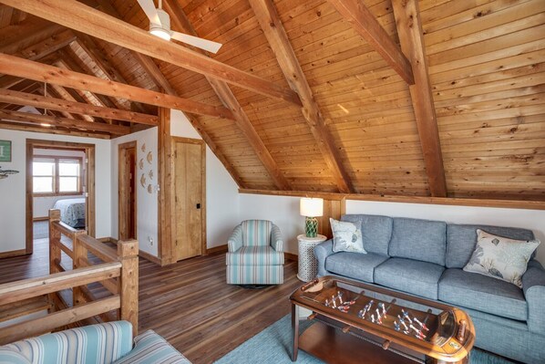 A cozy room with a wooden ceiling, a sofa, a striped armchair, a small table, and a bed with a wooden headboard. - Moon Rider: Oceanfront, Classic Style, Great Views (Nags Head)