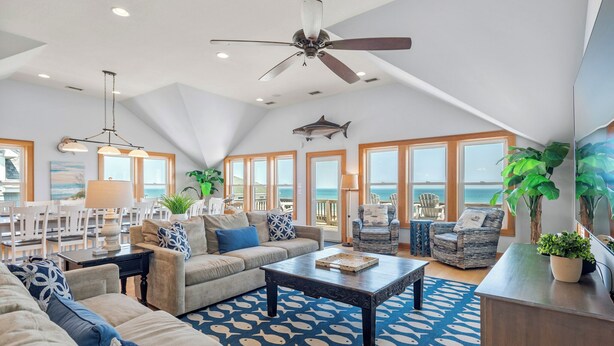 A spacious living room with a high ceiling, a ceiling fan, and a large window overlooking the ocean. There are multiple seating areas, including a sofa, chairs, and a dining table. - Cobia: Oceanfront Home w/ Private Heated Pool, Hot Tub & Cabana Service (Nags Head)