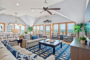 A spacious living room with a high ceiling, a ceiling fan, and a large window overlooking the ocean. There are multiple seating areas, including a sofa, chairs, and a dining table. - Cobia: Oceanfront Home w/ Private Heated Pool, Hot Tub & Cabana Service (Nags Head)