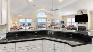A kitchen with a black countertop, white cabinets, and a ceiling fan. There is a dining area with chairs and a table, and a living room with a TV and seating. - Turtles Roost: Oceanfront, Pool, Spa, Game room (Nags Head)