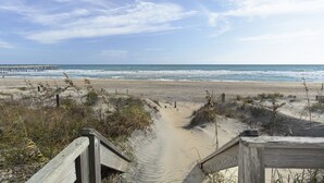 A beach with a wooden boardwalk leading to the water, sandy dunes, and a pier in the distance. - Turtles Roost: Oceanfront, Pool, Spa, Game room (Nags Head)