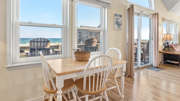 A dining area with a wooden table and white chairs, large windows with ocean views, and a door leading to a balcony. - Turtles Roost: Oceanfront, Pool, Spa, Game room (Nags Head)