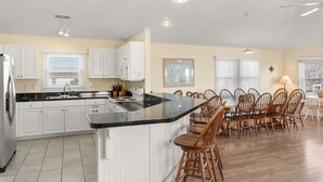 A modern kitchen with white cabinets, a black countertop, and wooden chairs. - Turtles Roost: Oceanfront, Pool, Spa, Game room (Nags Head)