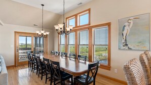 A dining room with a large wooden table, black chairs, a chandelier, and large windows with blinds. - Salt Shaker: Semi-oceanfront, Private Pool, Spa (Nags Head)