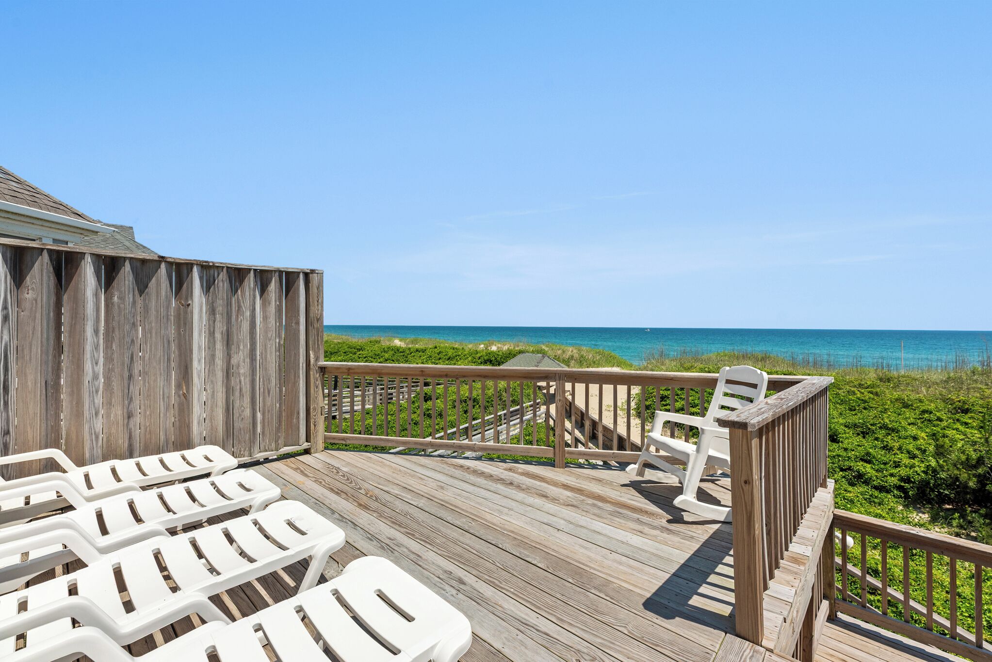 Une terrasse en bois, avec des chaises en plastique blanches et une rambarde en bois, donnant sur une plage.