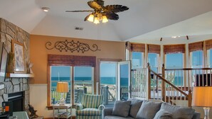 Ein geräumiges Wohnzimmer mit großem Fenster zum Meer, einem Kamin, einem Deckenventilator und einer Treppe, die zu einer Außenterrasse führt.