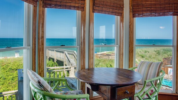 Ein Zimmer mit großen Fenstern mit Blick auf den Ozean, einem Holztisch und Korbstühlen.