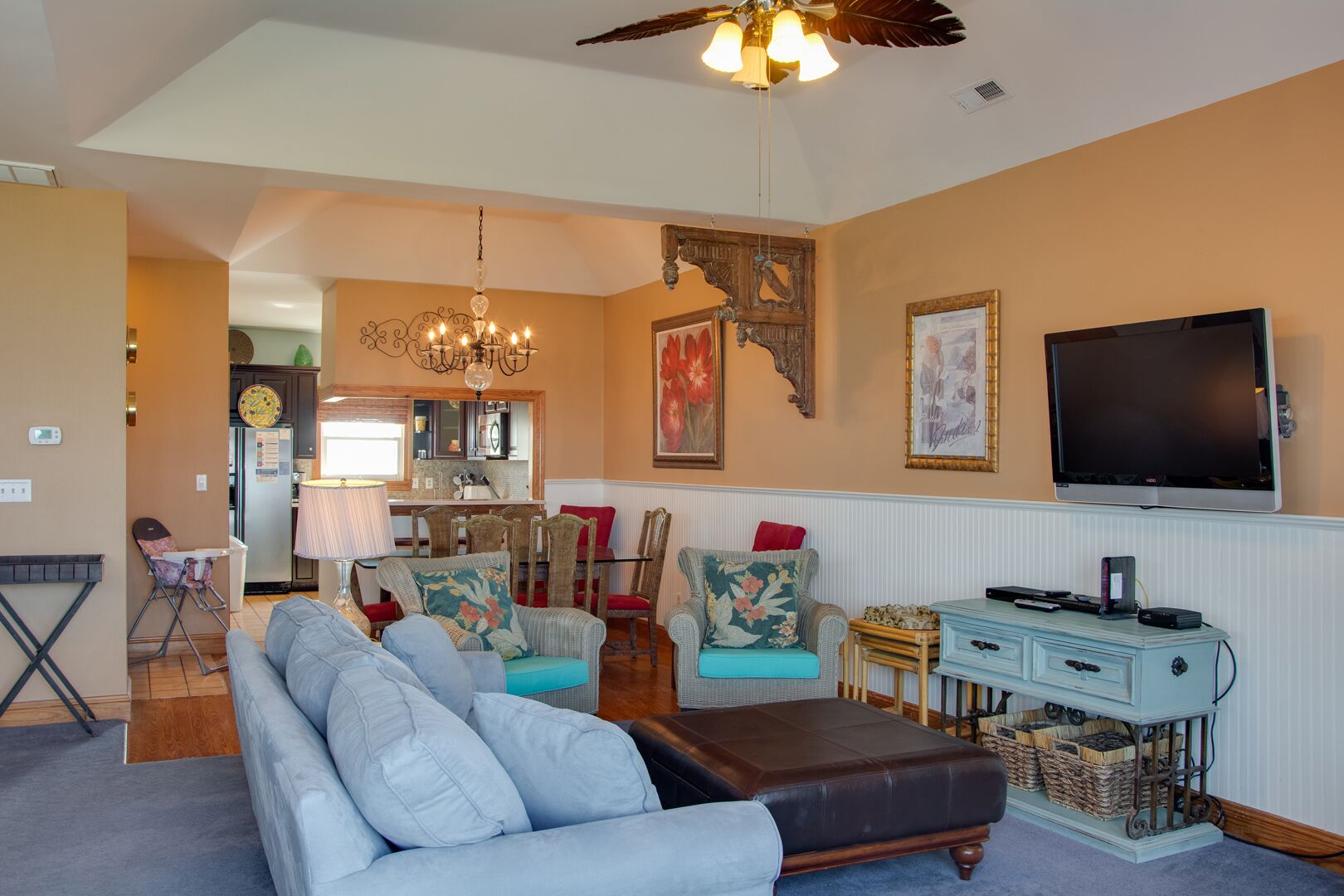 Una sala de estar con sofá, reposapiés y sillones, un comedor con mesa y sillas, una cocina con refrigerador y un reloj, y un televisor montado en la pared.