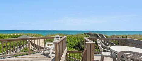 A wooden deck with a table and chairs, overlooking a beach with clear blue water and grassy dunes.