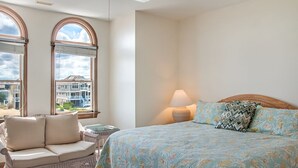 A bedroom with a bed, a wicker chair, a lamp, and large windows with a view of buildings. - Sea-Duced: Oceanfront, Great Views, Shared Pool. (Nags Head)