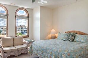 A bedroom with a bed, a wicker chair, a lamp, and large windows with a view of buildings.