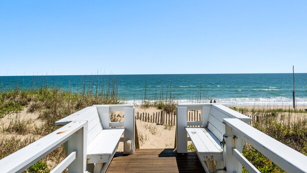 A wooden deck leading to a beach with white railings and benches.